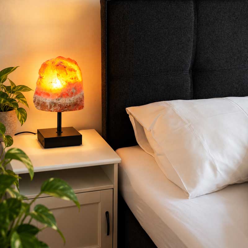 Aragoniet Edelsteen Lamp Golden Flame met warme goudgele en oranje verlichting op een wit nachtkastje, naast groene kamerplanten in een moderne slaapkamer.