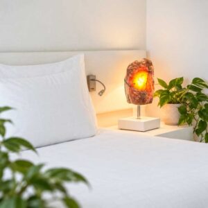 Aragoniet Edelsteen Lamp Lava Glow met warme oranjegele verlichting op een wit nachtkastje, naast een groene kamerplant in een moderne slaapkamer.