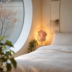 Roze Amethist Edelsteen Lamp Pink Moon met warme achtergrondverlichting op een wit nachtkastje, naast groene kamerplanten in een sfeervolle slaapkamer met rond raam.