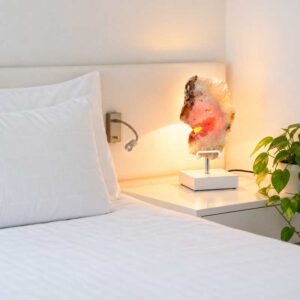 Roze Amethist Lamp Crystal Rose met witte standaard en warme LED-verlichting op een nachtkastje in een lichte slaapkamer met groene kamerplanten.
